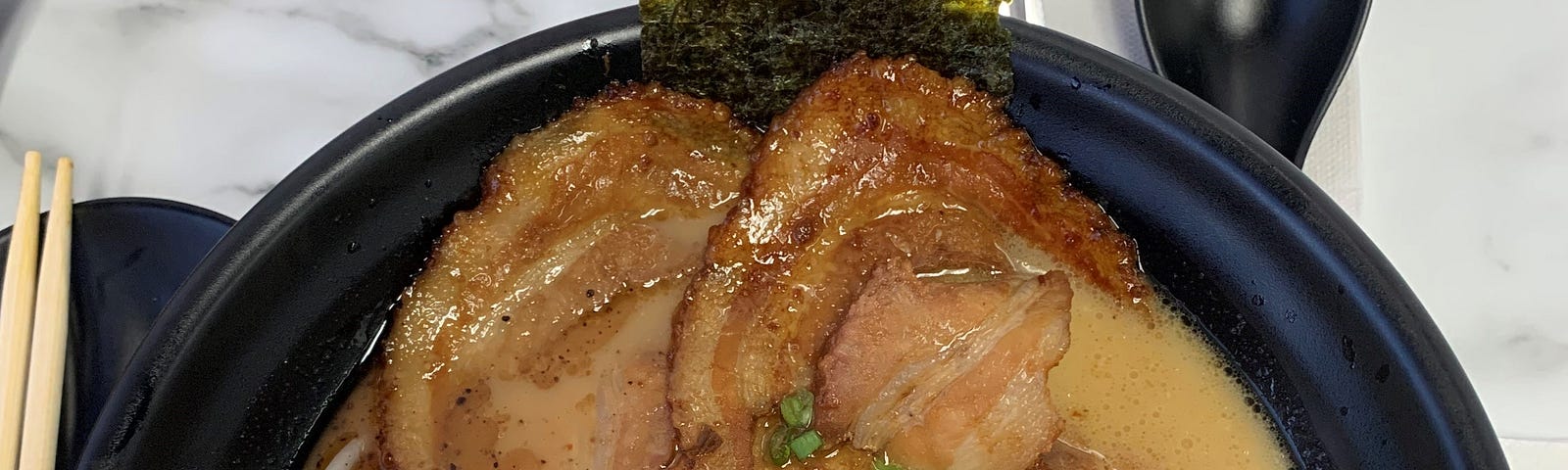A black bowl of ramen is topped with a fish cake, sprouts, green onion, bamboo shoots, and pork belly.