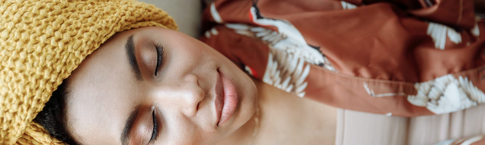 A woman with a headwrap on and a robe is laying on the couch with her eyes closed.