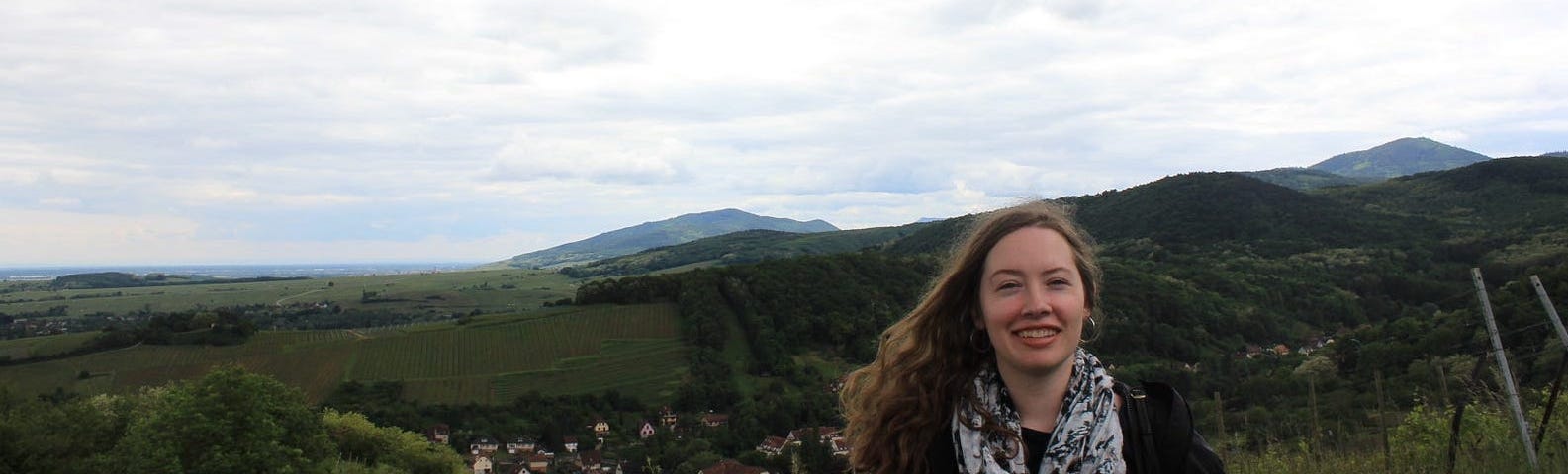 A tourist dressed in a wind jacket and floral scarf smiling in front of a backdrop of rolling hills, red-roofed houses and cloudy skies.