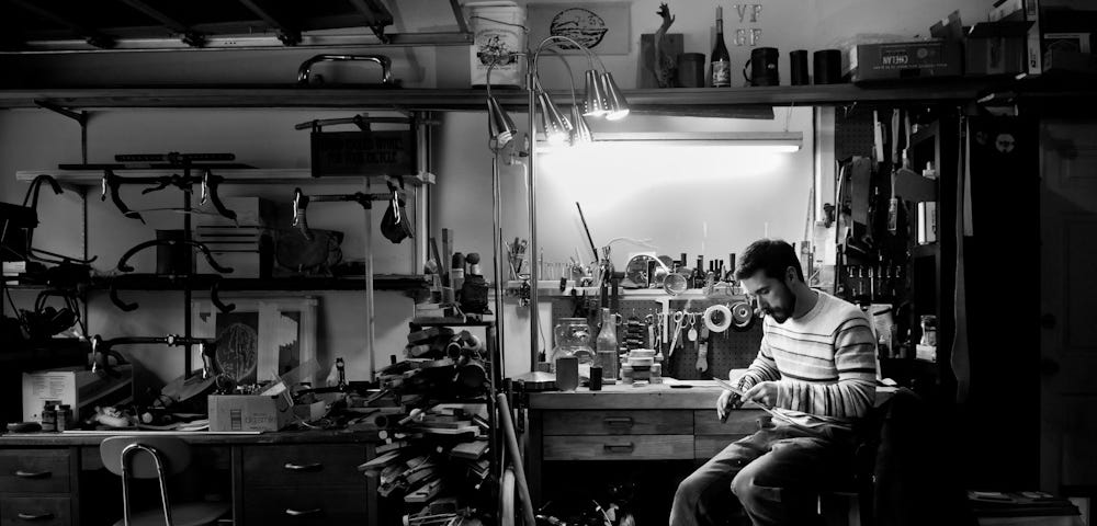 Black and white photo of a man working at a bench in a cluttered leather workshop in a garage. Over his workbench is a light and a window. Photo credit to Russ Roca / Path Less Pedaled.