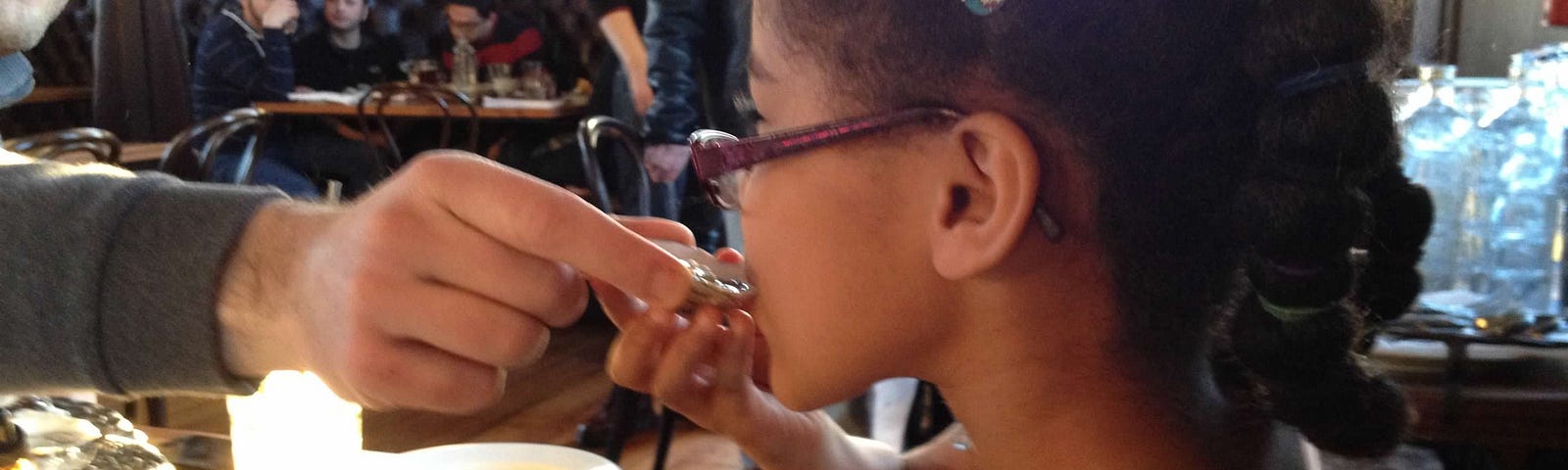 A young girl at a restaurant tries an oyster with help from her dad