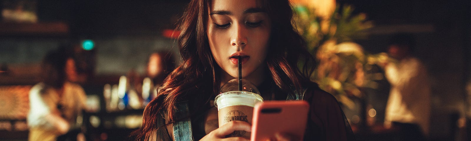 A young woman in a coffee shop looks at her phone while sipping a drink