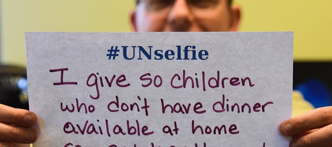 A man holding an #Unselfie sign. He handwrote on it: “I give so children who don’t have dinner available at home can eat healthy meals at afterschool programs.” The date is GivingTuesday, Dec 3, 2019