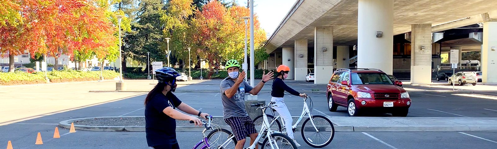 Three people straddle bikes in a sunny parking lot. Dan Hernandez (center) gestures while teaching the two class participants at an adult learn-to-ride class.
