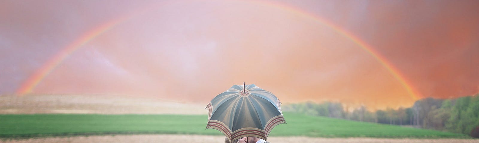 Couple kissing while holding an umbrella in a field with a rainbow in the background.