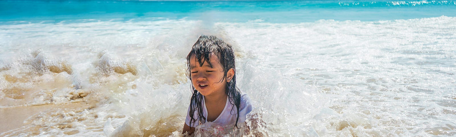 A young child sits on the sunny beach as waves splash all around them.