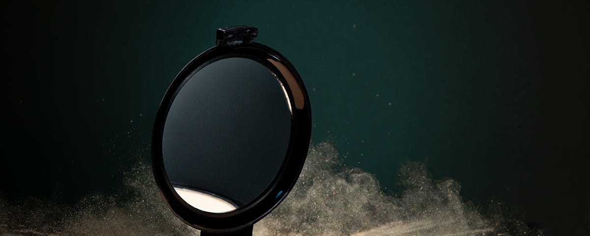 This is a horizontal photograph of an open cosmetic compact. We see the round mirror and the round base which holds the face powder. The compact was dropped onto a small pile of powder and the resulting image shows a frozen action photograph of the outwardly expanding powder explosion. Shot on a black plexiglass surface we also see powder rising in the air behind the compact.