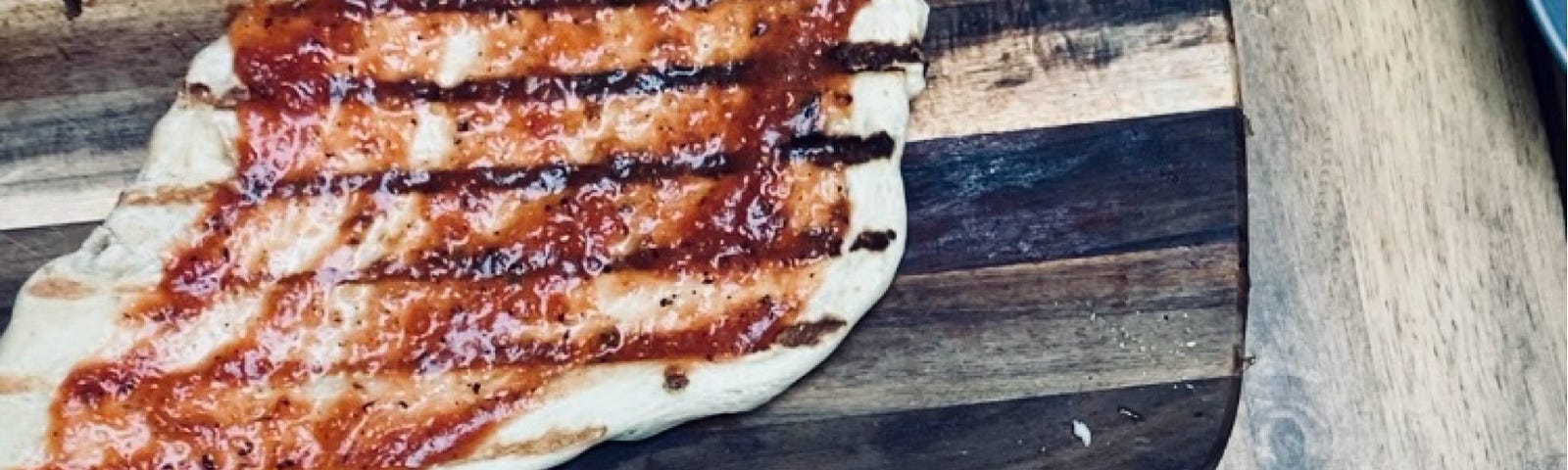 A photo of a grilled pizza in the making: a blob shaped dough with grill marks being spread with sauce, next to containers of toppings (pepperoni and mushroom)