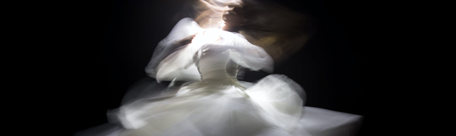 Photo of a woman in a white dress dancing in front of a black background. She’s twirling, her dress and scarves flaring out in all directions from her body. Her blonde hair covers her face as she continues to spin. A vertical white light shines on her head, like a connection with Spirit or God.