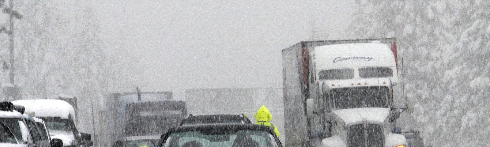 A person is kneeling in the middle of the road during a snowstorm to look at the tires of the car. There are skid marks and heavy traffic surrounding the car.