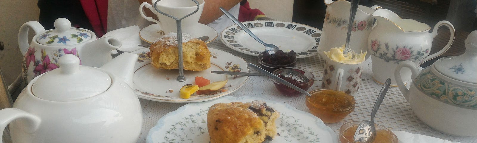 A table with lovely porcelain teapots, cups and saucers, with some scones, clotted cream and homemade jam.