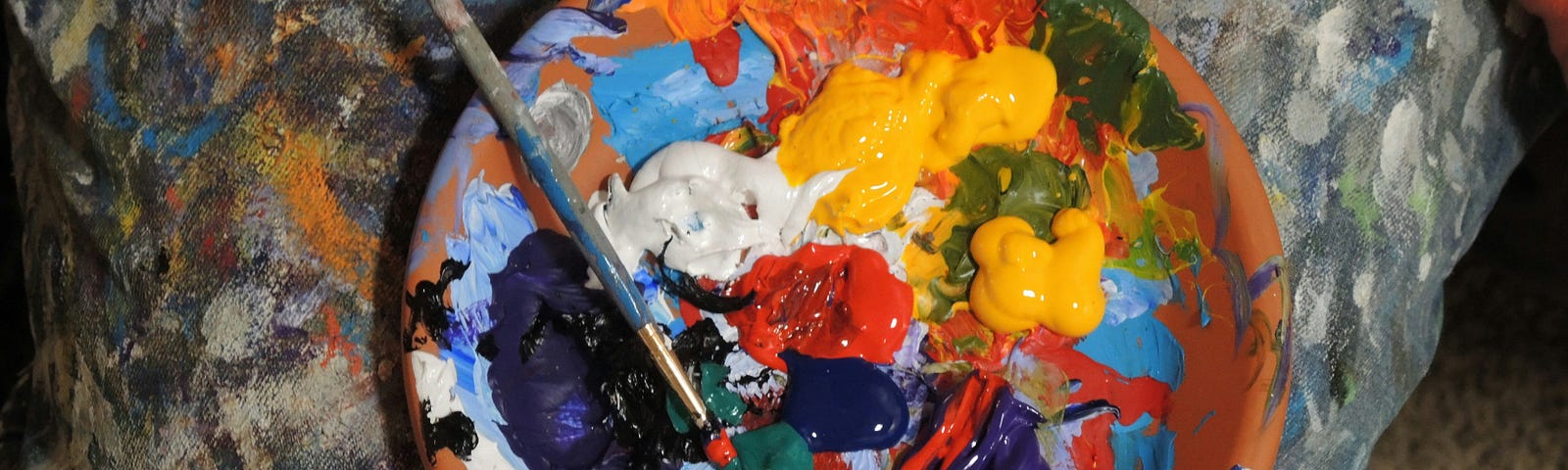 A painter palate with oil paints of yellow, blue, orange, red and green and other colors, in a wooden bowl with a paint brush dipped into the green.