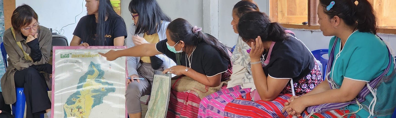 7 women are visible, they are wearing colourful traditional clothing and one of the women is pointing at a map.