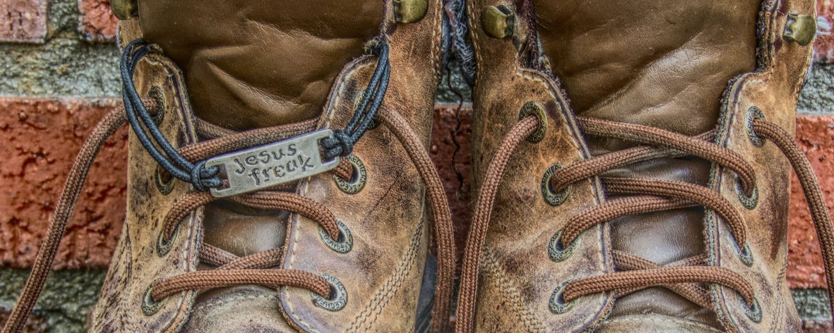 This image shows a pair of brown hiking boots with a silver tag that says “Jesus Freak” tied to the laces. This photograph is © Copyright 2010–2023 to Deb Wax, the photographer. All Rights Reserved.