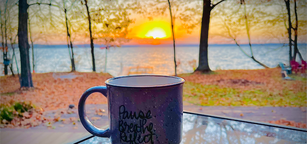 A tranquil lake-front morning sunrise, with a coffee mug on a glass table, reflecting the morning colors
