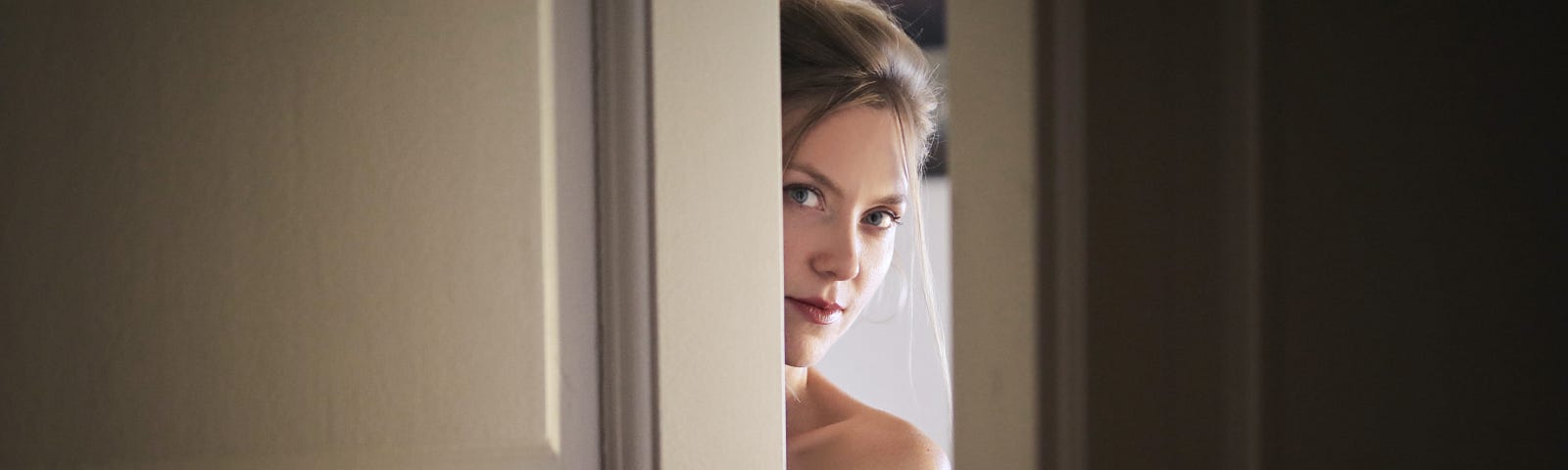 An attractive, young woman looking out through the crack of a partially open door.