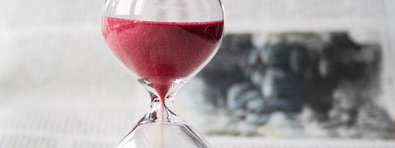 Close-up of an hourglass with red sand flowing through it, against a blurred newsprint background