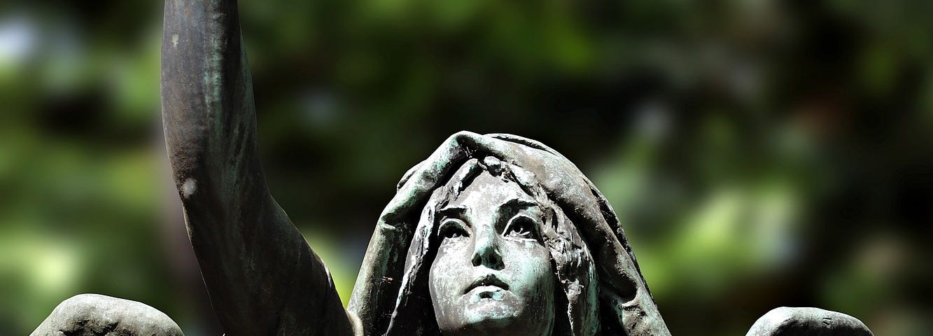 Statue of Angel looking up, right arm pointing upwards, cloth draping over her head and shoulders, wings seen from behind, greenery in backround.