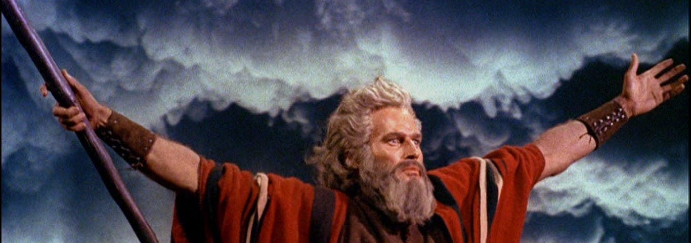 Color movie still of Charlton Heston as Moses in The Ten Commandments, 1956, with arms outstretched against a dark sky, parting the Red Sea.