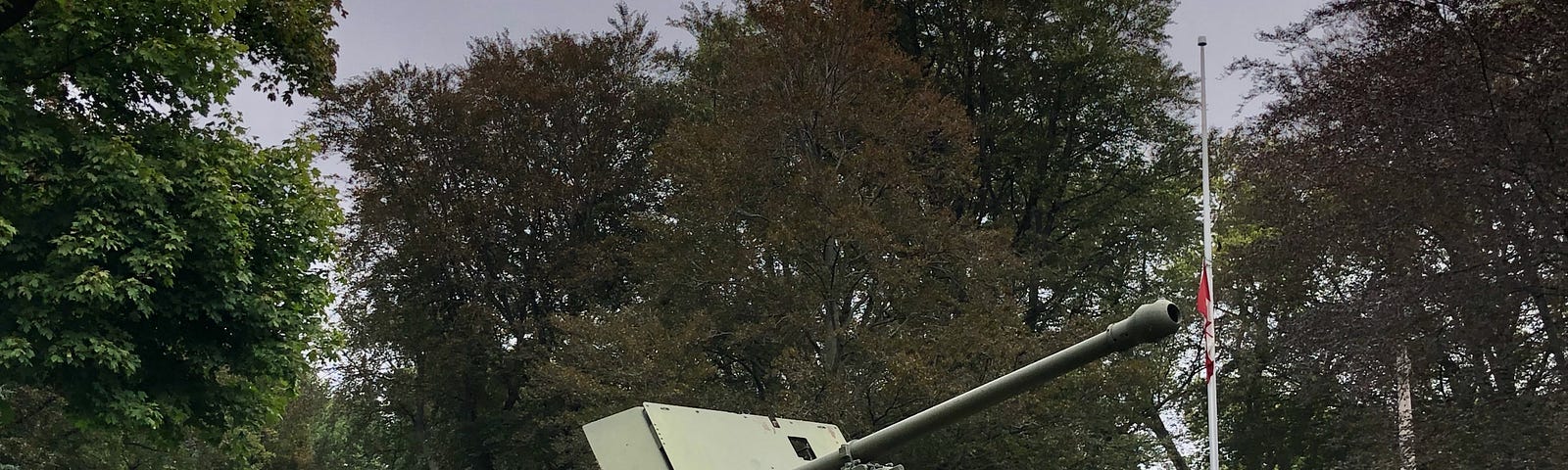 A gun is mounted on sand bags pointing off into the sky. It looks vintage as if it has been used in World War II. The grass is green around it and there are trees behind with a small amount of grey sky at the top of the photo