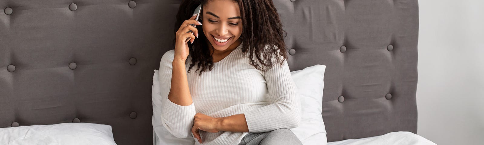 A happy Black woman sits on the bed with her laptop beside her. She smiles as she chats on the phone.