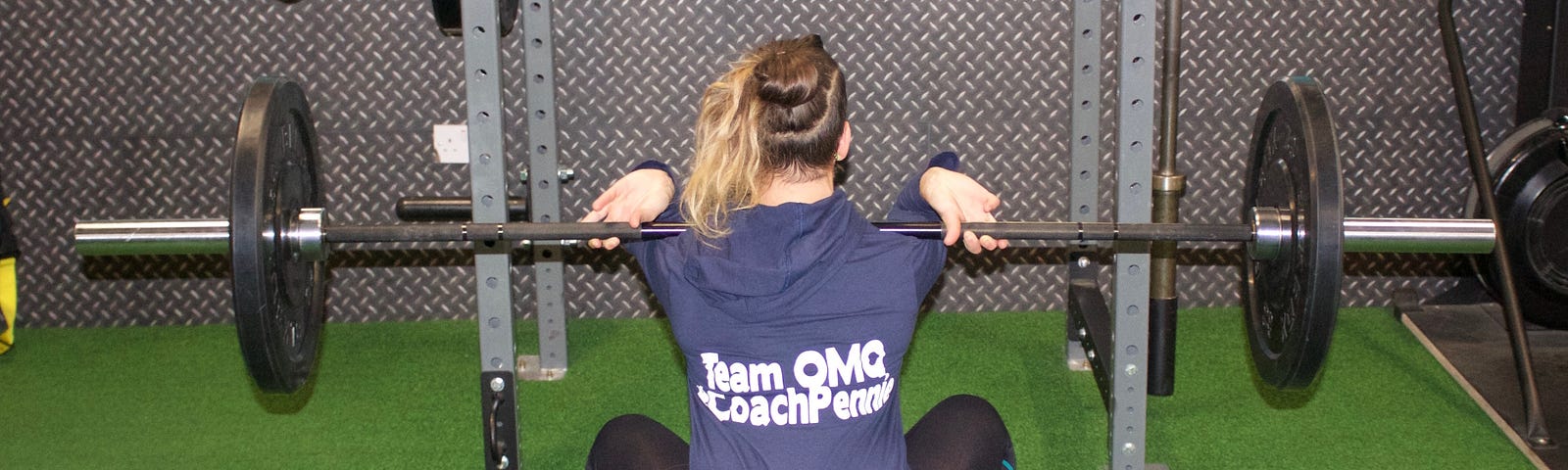 Photo shows Pennie Varvarides, author of the article and strength coach, performing a barbell front squat. Pennie is wearing pink crossfit nanos, black leggings and a blue top which reads “Coach Pennie”.