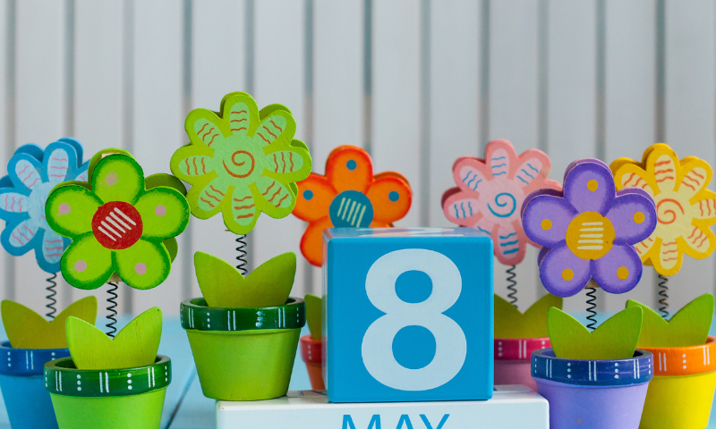 A colorful photo of plastic flowers in matching pots sitting on a pastel blue table next to a white rectangular block that says “May.” On top of this block sits a medium blue shaded cube with a white “8” for today’s date. The flowers are in assorted colors, lime green, pink, yellow, purple, and blue. The background is a white wall with grey pinstripes. The “8” in the photo also represents the Monday Morning Huddles' official morning timeslot of 8 AM. Get ready for progress and abundance!