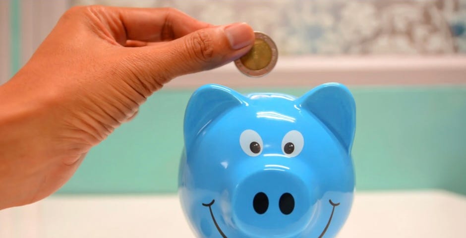 A hand dropping a coin into a piggybank; you can also save money as a writer by getting free writing lessons