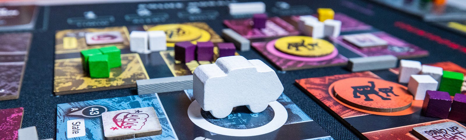 A corner of the Bloc by Bloc: Uprising board. A white police van meeple sits in a white ring on the Liberated Telecom Network Hub district tile, along with 5 white police squad cubes. A graffiti token covers a shop icon on the tile. Adjacent and kitty corner to the tile are other liberated district tiles with an assortment of bloc and police cubes, graffiti tokens, and barricades in between.