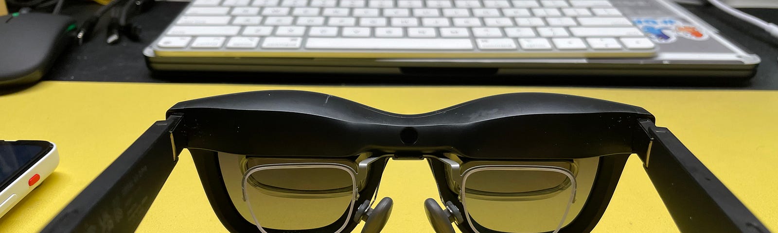 Augmented reality glasses on a yellow desk, with a keyboard and other electronic devices in the background.