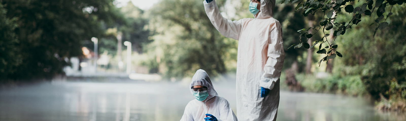 Two researchers in protective suits and masks collecting water samples from a river for glyphosate analysis