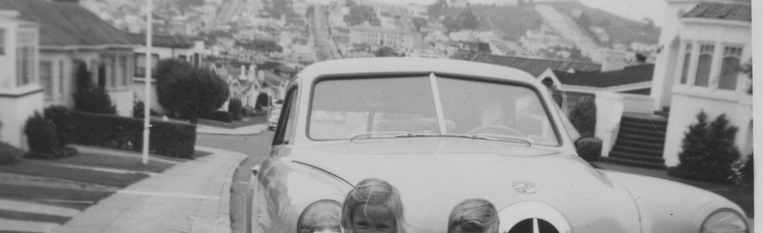 Two small children stand in front of a car parked at a curb facing uphill. Portions of houses can be seen down the hill behind the car with another hill in the background that is covered with buildings.