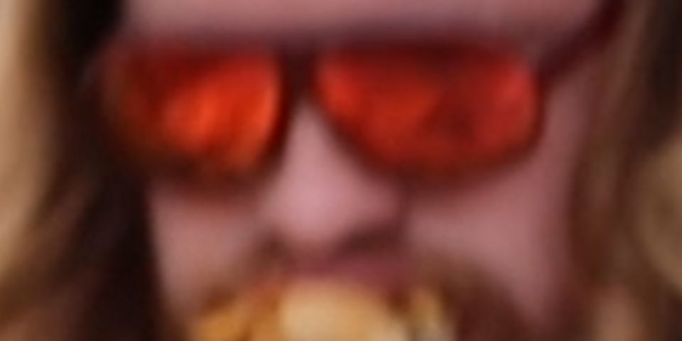 Blurry image of a man violently stuffing a second cheeseburger into his mouth which is already full with a first cheeseburger.