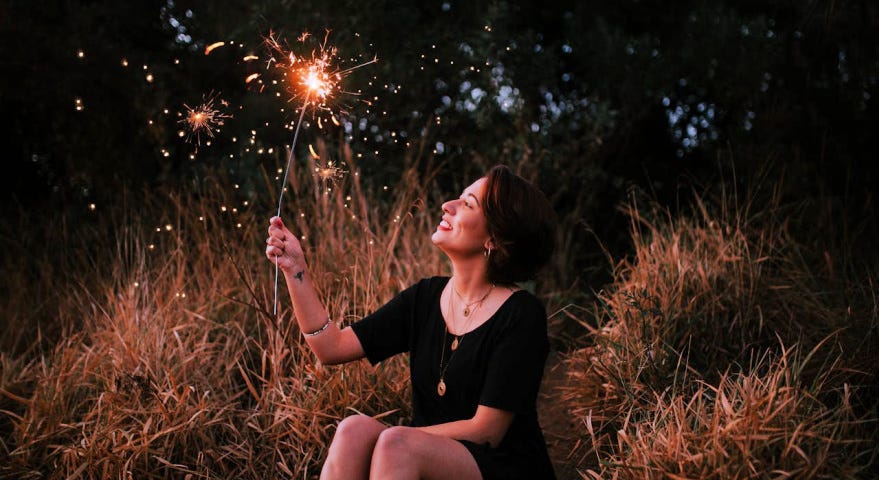 A dark-haired woman sitting on a hillside, looking up at the sparkler she’s holding in her hand.