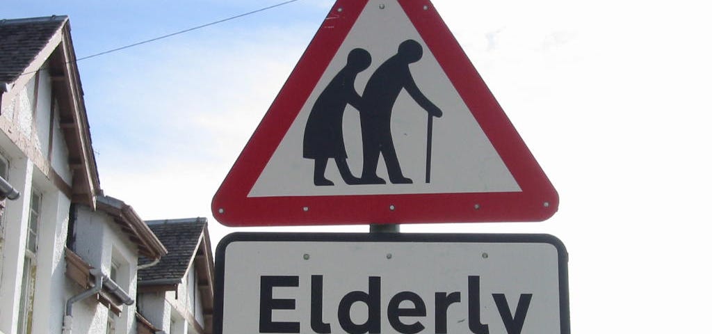 A British road sign, a red triangle with two people hunched over using walking sticks, with a rectangular sign below with the words, “Elderly people”.