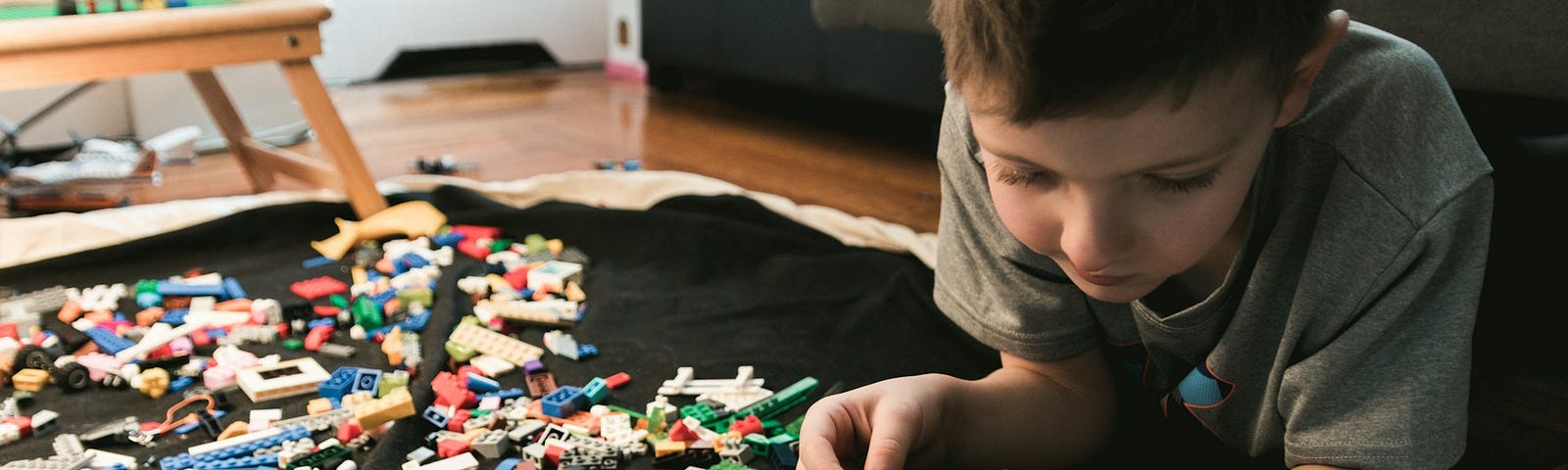 A young boy is lying on his belly in a living room, playing with Lego and looking at a book of Lego designs. The boy has short brown hair and is wearing a grey T-shirt.