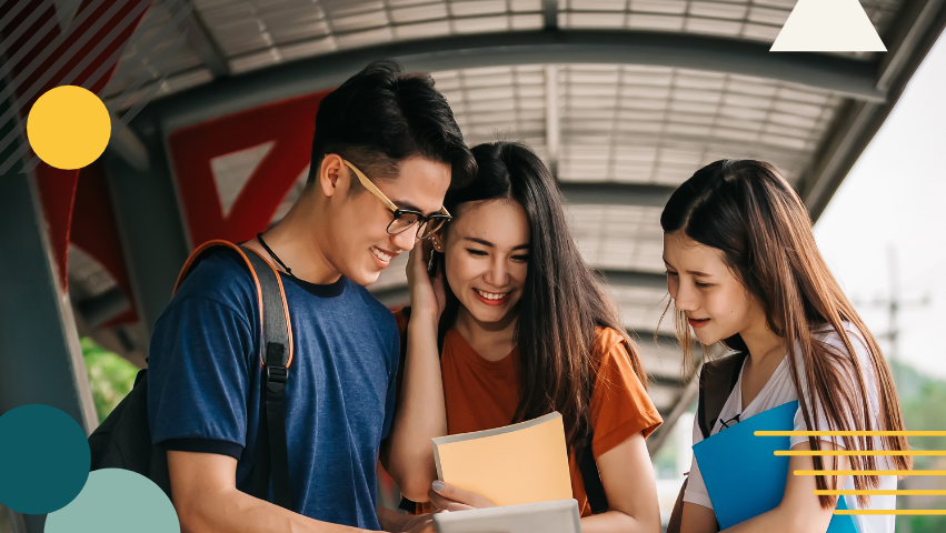 Three middle or high school aged students huddle together and look at a notebook while smiling.