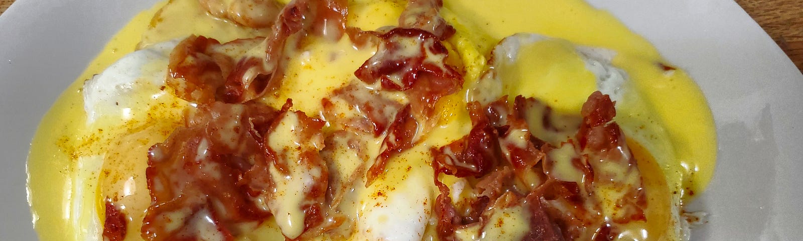 A white square plate with three potato patties with one fried egg on each and all topped with crisp prosciutto and homemade hollandaise sauce.