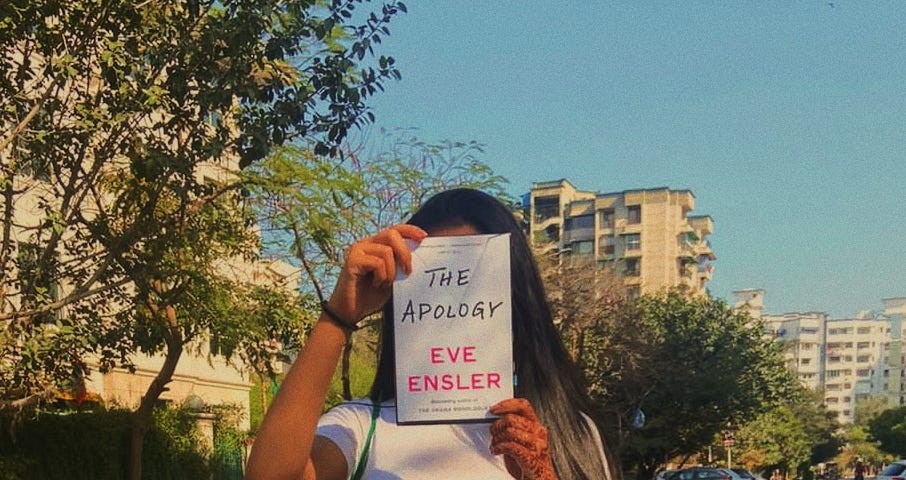 The author holding up a paperback of ‘The Apology’ by Eve Ensler on a sunny day within a city landscape.