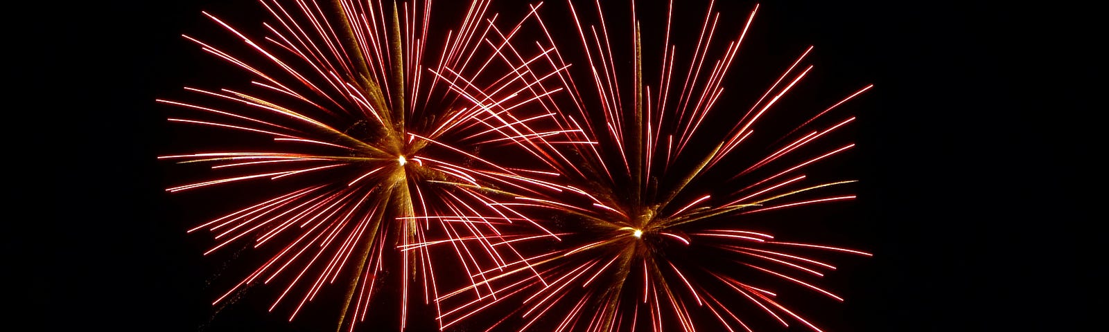 Four firework shells exploding in the sky in dandelion fluff shape. The two largest are pink and gold, the two smaller ones just below are gold.