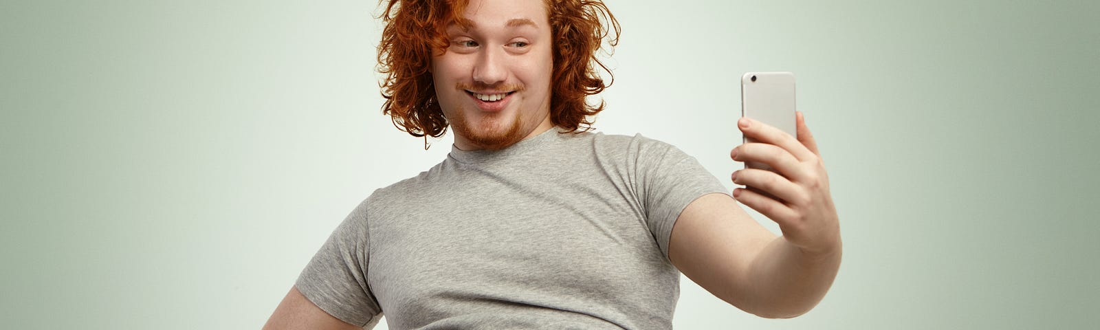obese-young-male-with-curly-ginger-hair-beard-holding-mobile-phone-posing-selfie-looking-with-flirty-smile-while-his-fat-belly-hanging-out-grey-shrunk-t-shirt-jeans-pants — Un Swede