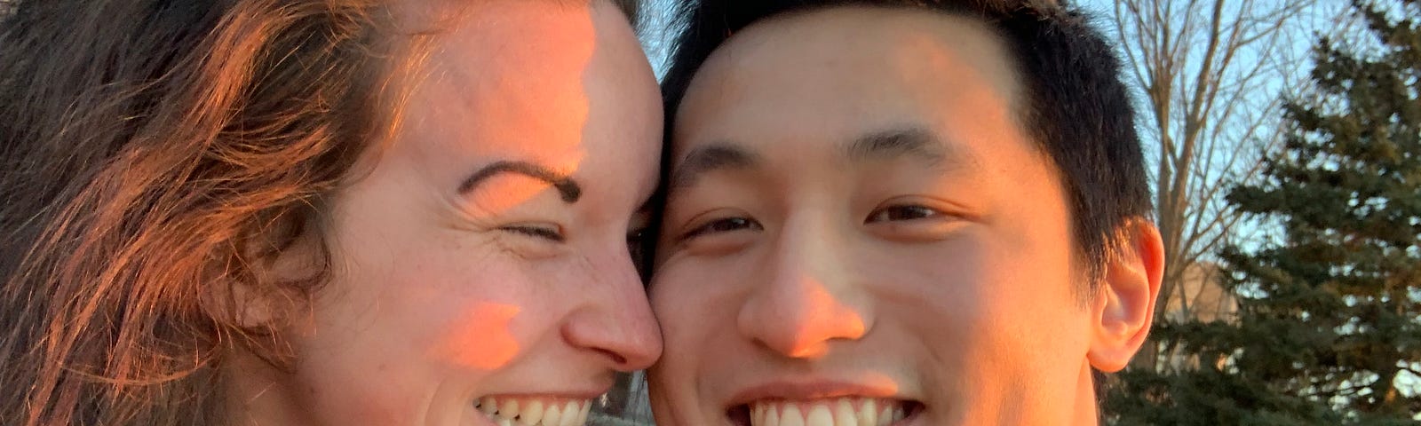 My husband and I sitting on a bench on campus. I (a white female with long curly brown hair and regrettably done eyebrows) am sitting on the left wearing a red and black flannel with a black denim jacket. Taka (an East Asian guy with a cheesy smile) is sitting on the right wearing a green and black flannel with a bright yellow jacket. We are both smiling and I am leaning into him. There are pine trees in the background.