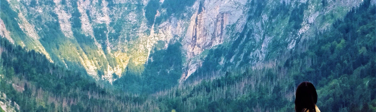 girl with long dark hair wearing yellow dress sitting on boulder looking out over lake and mountains