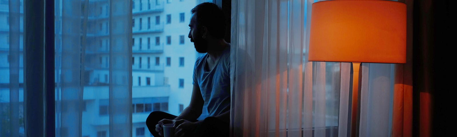 man sitting on window ledge in darkened hotel room with lamp