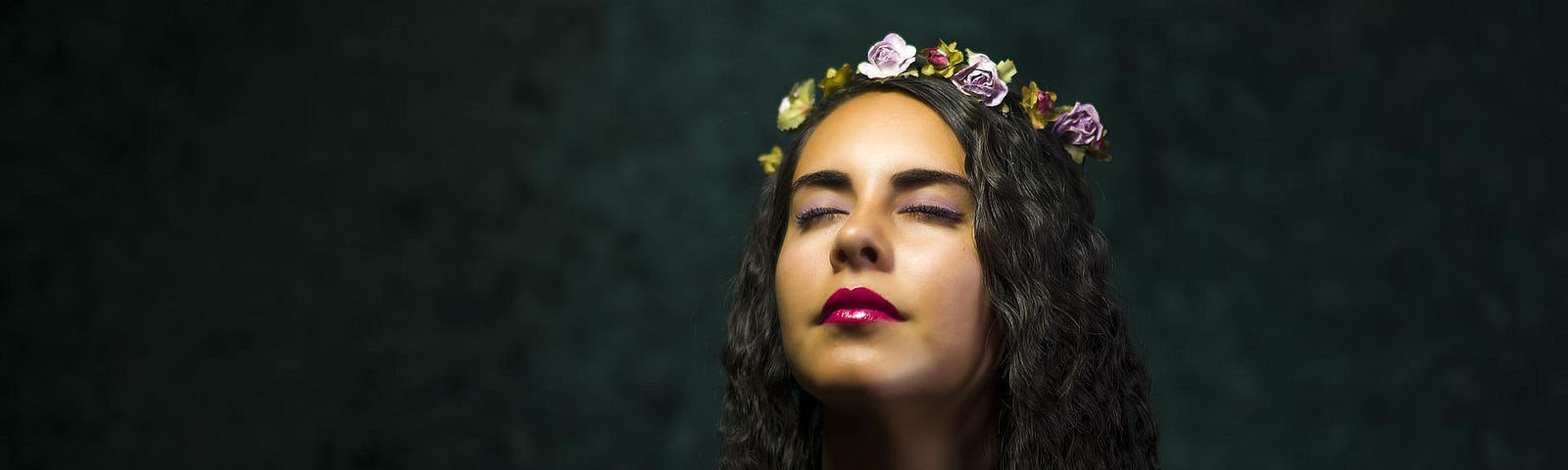 A woman with light brown skin and shoulder length black hair with her head tipped back and her eyes closed. She is wearing a crown of pale purple flowers, red lipstick, and a dark green shirt. The background is blurred and dark.