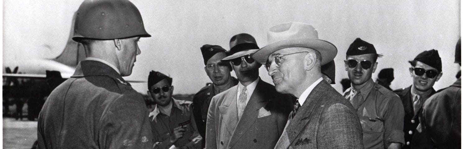 President Harry Truman stands in front of a group of soldiers on an airplane tarmac. He wears a brown suit, a white hat, and has his hands in his suit pockets. His head is turned to look at a soldier in a uniform and helmet, whose face we see in profile.