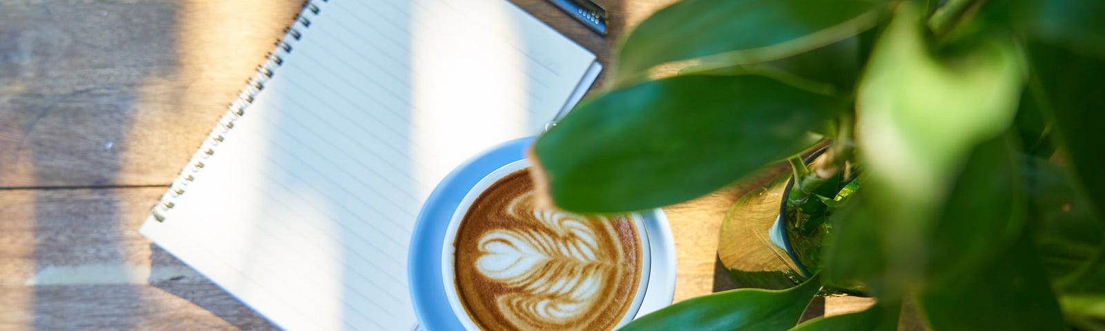 Imagem encontrada no site Pexels, nela podemos ver uma superfície marrom que remete a madeira, sobre ela há um bloco de notas branco, com uma caneta preta ao lado e uma xícara branca com café. Em primeiro plano da imagem há uma planta que sobrepõem os outros objetos da foto.