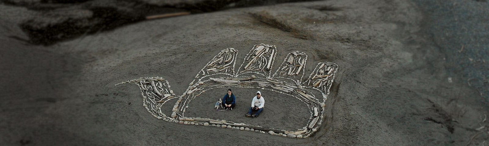 A massive Gitxsan formline artwork of an open hand made of driftwood and stones on a sandy riverbank. Two people and a dog sit inside the palm of the hand.