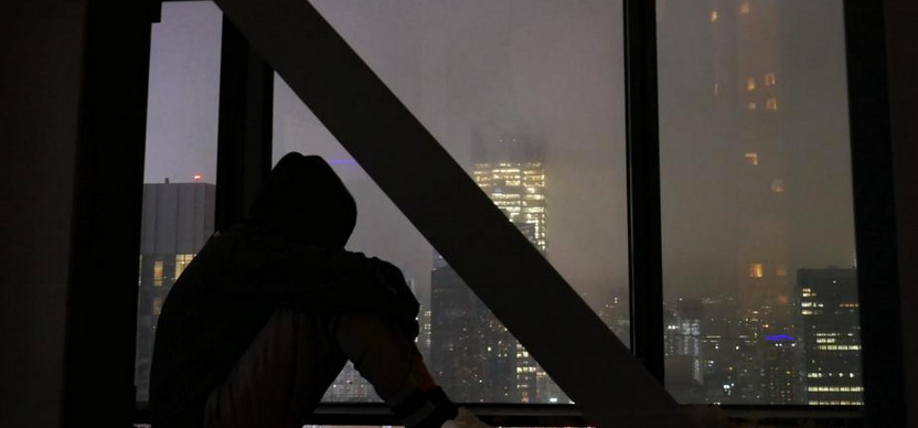 a hooded figure looks out a window at the New York night sky