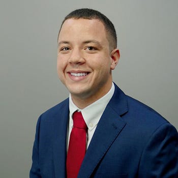 Trey Jackson is pictured wearing a navy blazer and a red tie against a gray background